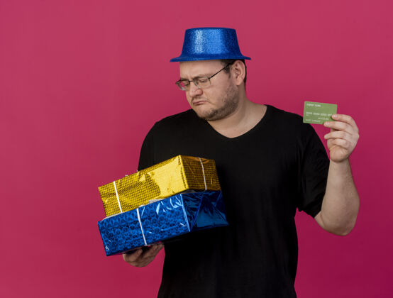 男士失望的成年斯拉夫人戴着眼镜 戴着蓝色派对帽 手里拿着礼品盒和信用卡蓝色粉色信用卡