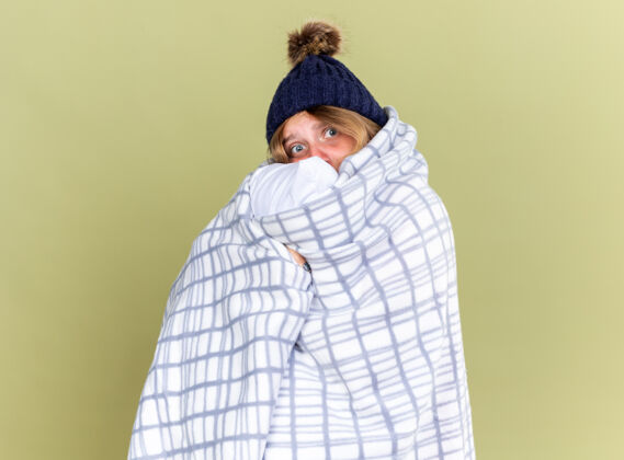 感觉不健康的年轻女子戴着暖和的帽子 裹着毯子 抱着枕头 站在绿色的墙壁上感觉不舒服枕头站着寒冷