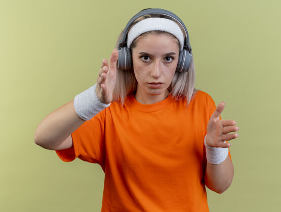 头带自信的年轻白人运动女孩戴着耳机戴着背带戴着头带和腕带双手直指镜头耳机直手