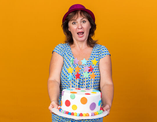 立场中年妇女戴着派对帽 手拿生日蛋糕 站在橘色的墙上 开心又惊喜地庆祝生日派对生日中间庆祝