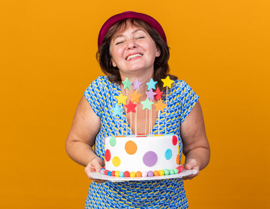 生日中年妇女戴着派对帽 手拿生日蛋糕 面带笑容 站在橘色的墙上欢庆生日兴奋庆祝举行