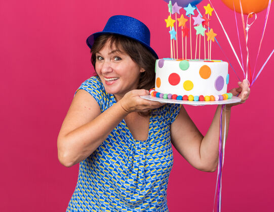帽子快乐快乐的中年妇女戴着派对帽 手拿彩色气球 手拿生日蛋糕 面带微笑 站在粉色的墙上庆祝生日派对中间生日气球