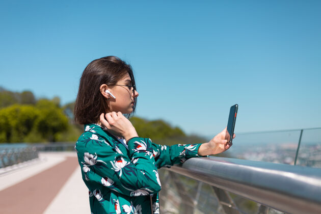 青少年阳光明媚的日子里 穿着休闲绿衬衫的女人站在桥上 看着手机屏幕 耳朵里戴着无线蓝牙耳机城市女性媒体