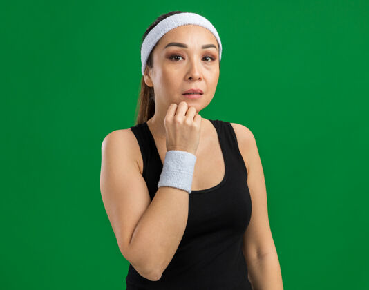 健身戴着头巾和臂章的年轻健身女士 神情沉思 手放在下巴上思考 站在绿色的墙上年轻表情思考