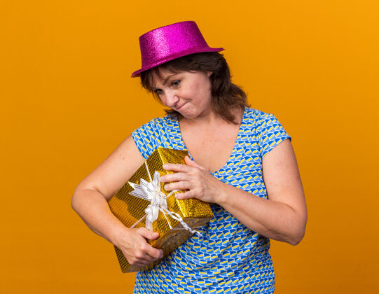 开放中年妇女戴着派对帽 手里拿着礼物 看起来很困惑 正准备打开盒子 站在橙色的墙上庆祝生日派对困惑年龄中间