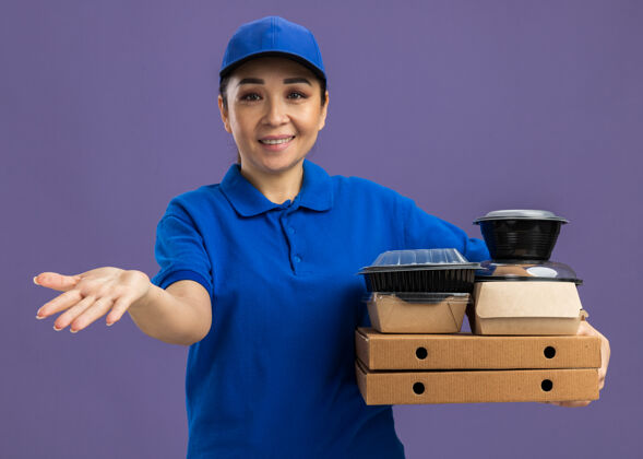 欢呼身穿蓝色制服 头戴鸭舌帽的年轻女送货员手举比萨饼盒和食品包 手伸微笑着站在紫色的墙上送货站着胳膊