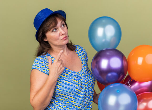 生日中年妇女戴着派对帽 手里拿着一堆五颜六色的气球 站在绿色的墙上 困惑地看着生日派对庆祝中间年龄