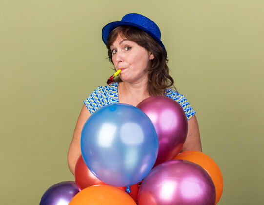 生日快乐而积极的中年妇女戴着派对帽 手里拿着一堆五颜六色的气球吹着哨子 站在绿色的墙上庆祝生日派对哨子正面年龄