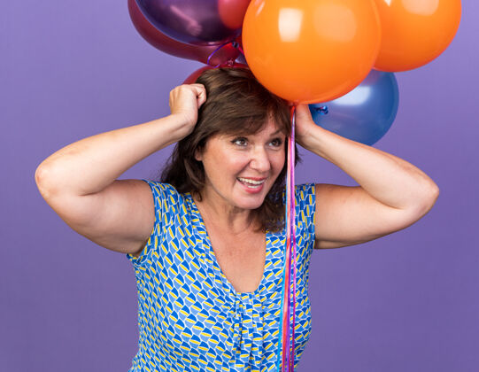 年龄快乐而兴奋的中年妇女手持一束五颜六色的气球站在紫色的墙上庆祝生日派对气球五颜六色立场