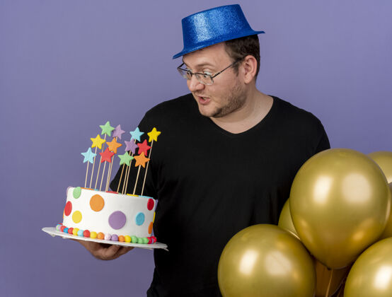 站立一个戴着眼镜 戴着蓝色派对帽的成年斯拉夫人惊讶地看着站着氦气球的生日蛋糕斯拉夫惊喜派对