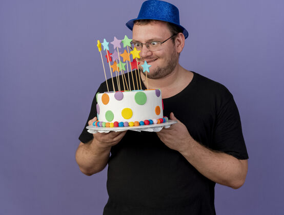 生日兴奋的成年斯拉夫人戴着眼镜 戴着蓝色派对帽 拿着生日蛋糕看光学男人帽子
