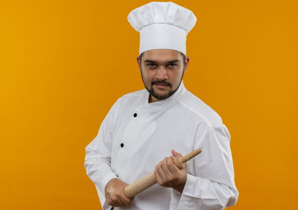 烹饪自信的年轻男性厨师穿着厨师制服拿着擀面杖 在橙色的墙上看起来很孤立 有复制空间持有信心卷