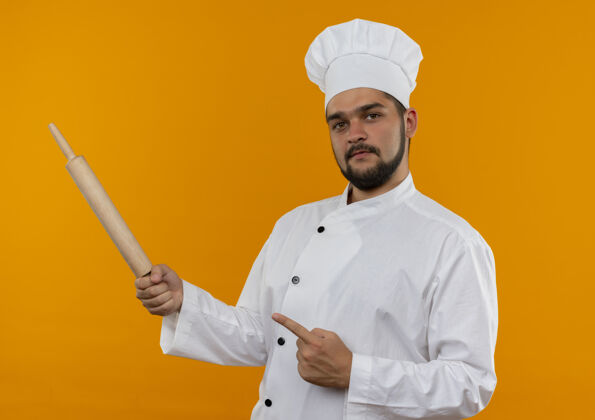 拿着自信的年轻男厨师穿着厨师制服 拿着擀面杖 指着橙色墙上孤立的一面烹饪制服别针