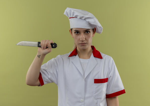 刀自信的年轻漂亮厨师穿着厨师制服 拿着隔离在绿墙上的刀持枪烹饪厨师