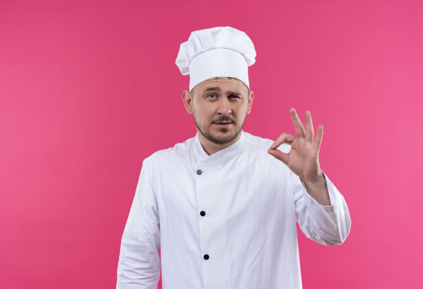 自信自信的年轻帅哥厨师身着厨师制服在粉红色的墙上做着“ok”的招牌年轻好的帅气
