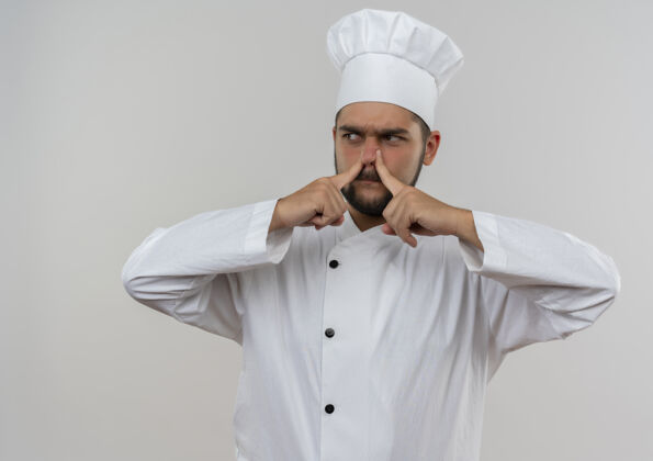 鼻子恼怒的年轻男厨师穿着厨师制服把手指放在鼻子上看着隔离在白墙上的一边男性烹饪恼火
