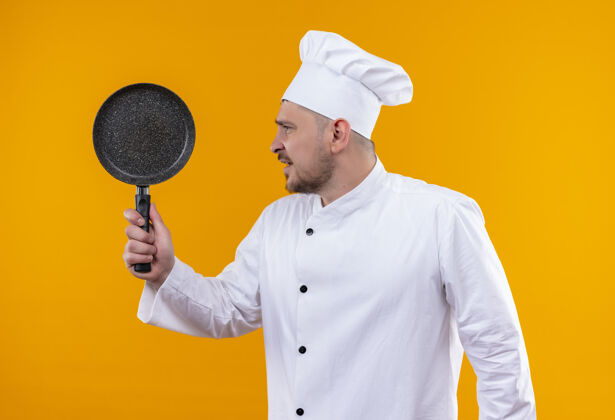 年轻年轻帅气的厨师穿着厨师制服 手里拿着煎锅 看着橘色墙上孤立的一面壁板薯条烹饪