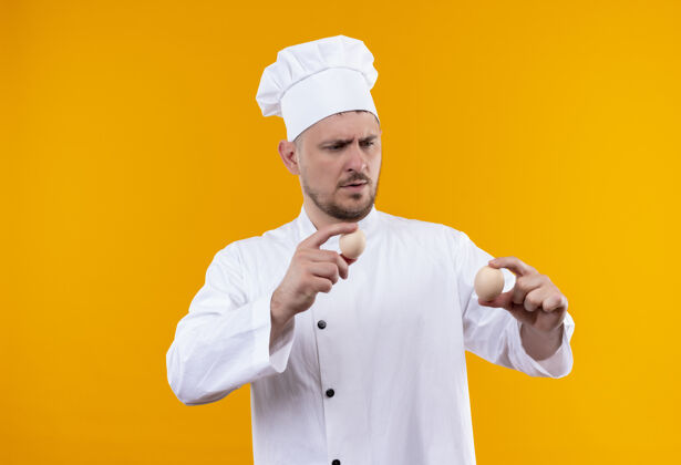年轻迷茫的年轻帅哥厨师穿着厨师制服拿着鸡蛋看着隔离在橙色墙上厨师持有鸡蛋