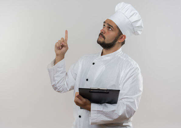 厨师穿着厨师制服的年轻男厨师手拿着剪贴板 孤立地指着白色墙壁上的复制空间年轻制服剪贴板