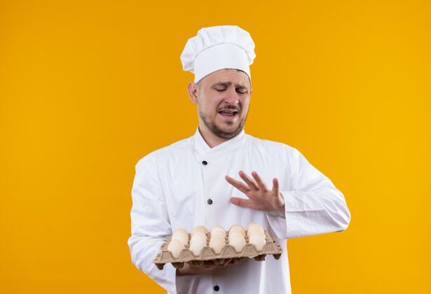 拿着恼怒的年轻帅哥厨师身着厨师制服 手里拿着一盒鸡蛋 在橙色的墙上做着不孤立的手势烹饪恼火帅气