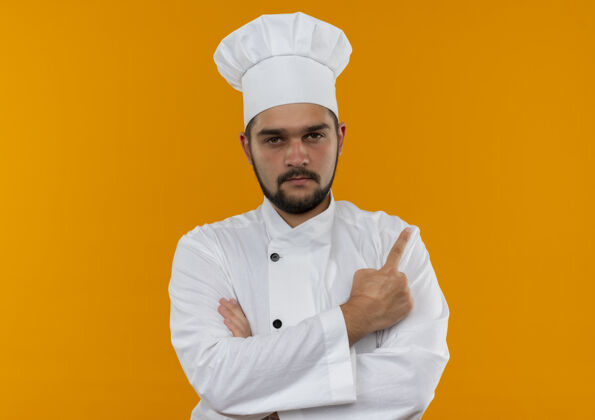指点自信的年轻男厨师身着厨师制服 站在封闭的姿势 指着橙色墙上孤立的一侧侧线封闭站立