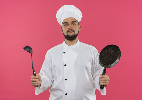 厨师自信的年轻男厨师身着厨师制服 手拿勺子和煎锅 隔离在粉红色的墙上年轻制服勺子