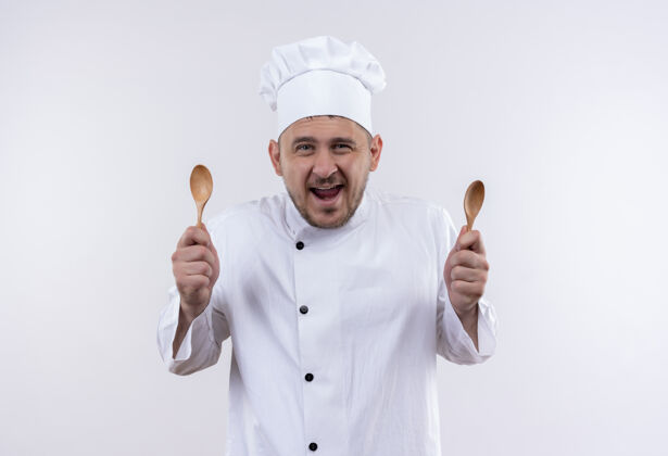 厨师快乐的年轻帅哥厨师穿着厨师制服拿着勺子站在孤零零的白墙上手持欢乐烹饪