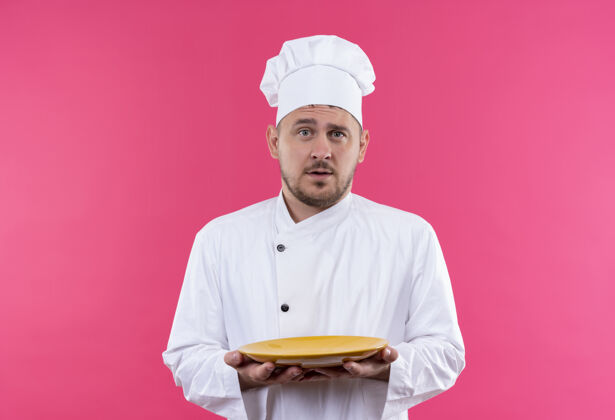手持印象深刻的年轻帅哥厨师在厨师制服举行盘子隔离在粉红色的墙上盘子帅气年轻