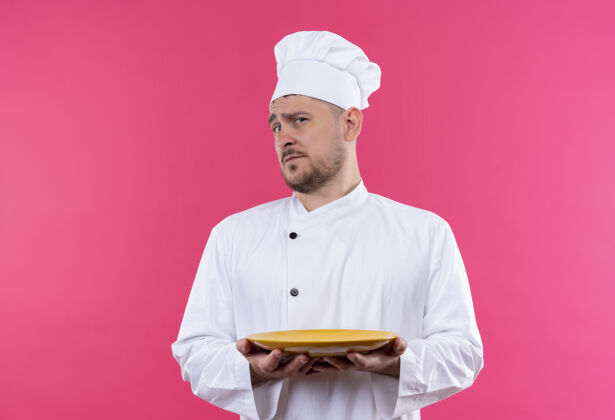 年轻迷茫的年轻帅哥厨师穿着厨师制服拿着盘子隔离在粉红色的墙上迷茫制服烹饪