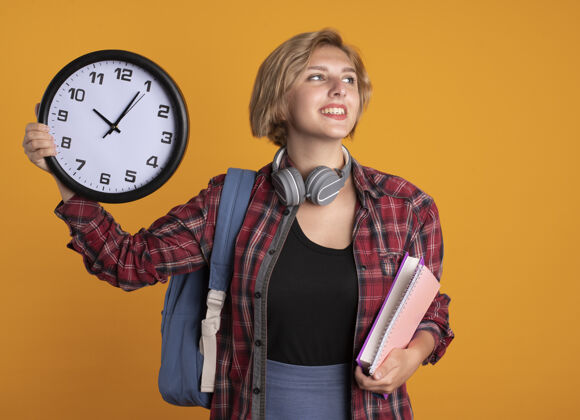 女孩面带微笑的年轻斯拉夫女学生戴着耳机 背着背包 拿着钟表本和笔记本看着身边空间背包耳机