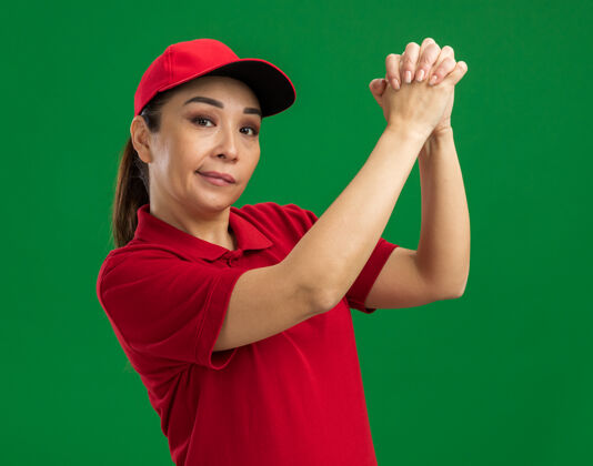 使身着红色制服 头戴鸭舌帽的年轻女送货员手拉手 站在绿色的墙边 自信地做着团队合作的手势交付年轻女人