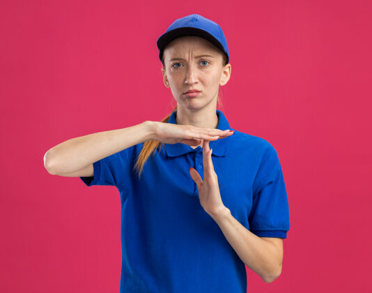 年轻身穿蓝色制服 头戴鸭舌帽 表情严肃的年轻送货女 双手站在粉色墙壁上做着暂停的手势制服递送帽子