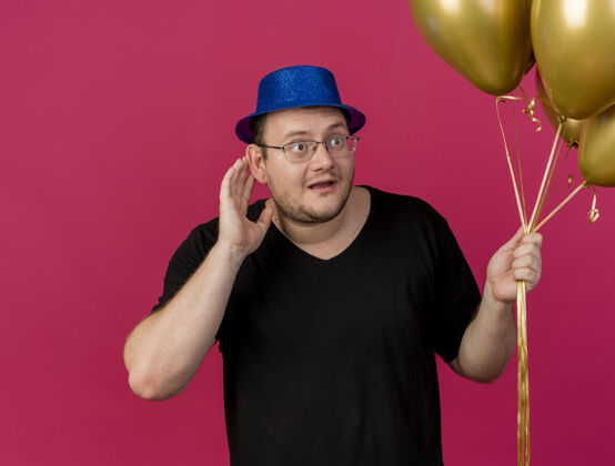 聚会令人印象深刻的成年斯拉夫人戴着眼镜 戴着蓝色的聚会帽 手贴着耳朵 试图听到并拿着氦气球保持空格氦