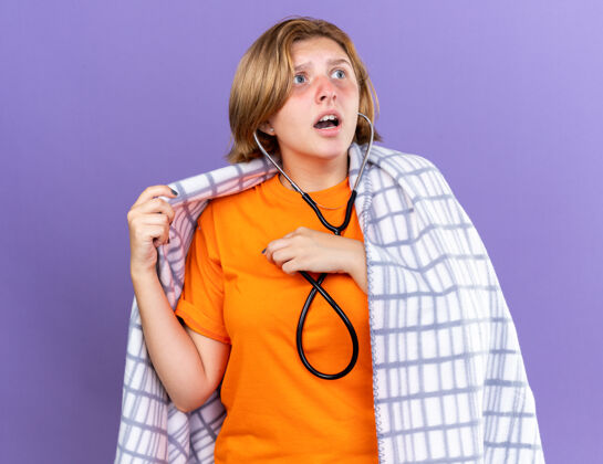 听诊器不健康的年轻女子裹着温暖的毯子 感觉不舒服 站在紫色的墙上 用听诊器听着自己的心跳 看起来很担心包装年轻毯子