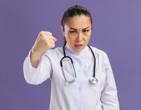 女人身穿白色药衣 脖子上戴着听诊器的年轻女医生站在紫色的墙壁上怒气冲天秀药医生