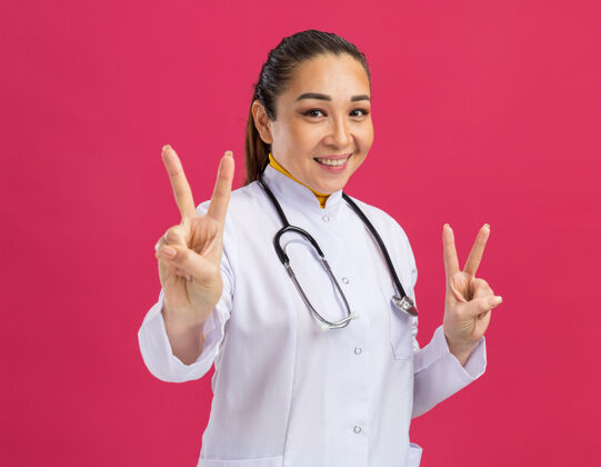 年轻人身穿白色药衣 脖子上戴着听诊器的年轻女医生微笑着站在粉色墙壁上的v形标志微笑外套医生