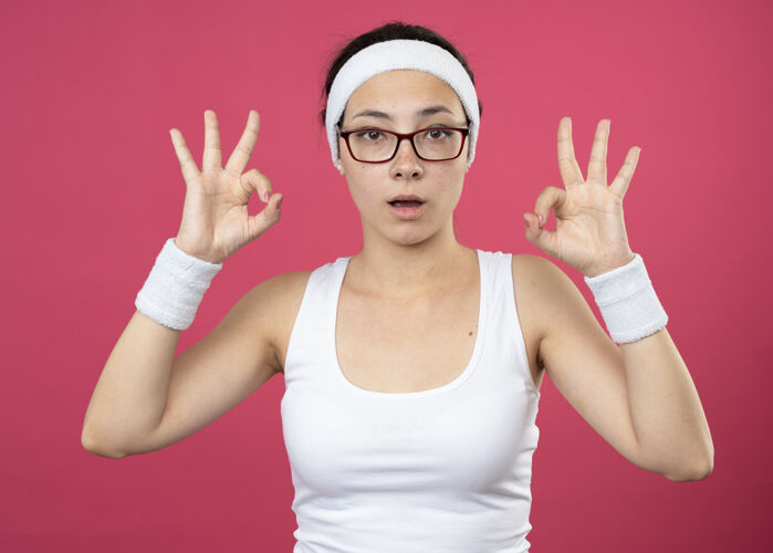 手势惊讶的年轻运动女孩戴着眼镜戴着头带和腕带手势ok手势用两只手佩戴标志腕带