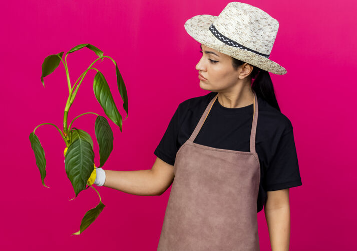 女人年轻漂亮的女园丁戴着橡胶手套围裙戴着帽子拿着植物严肃地站着看着它脸手套漂亮