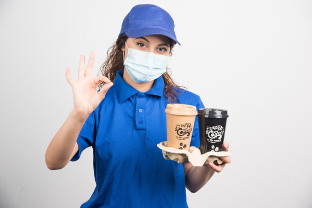 一个穿着蓝色制服 戴着医用口罩的女人 手里拿着两杯咖啡 大拇指朝着白色的脸手势年轻人成人