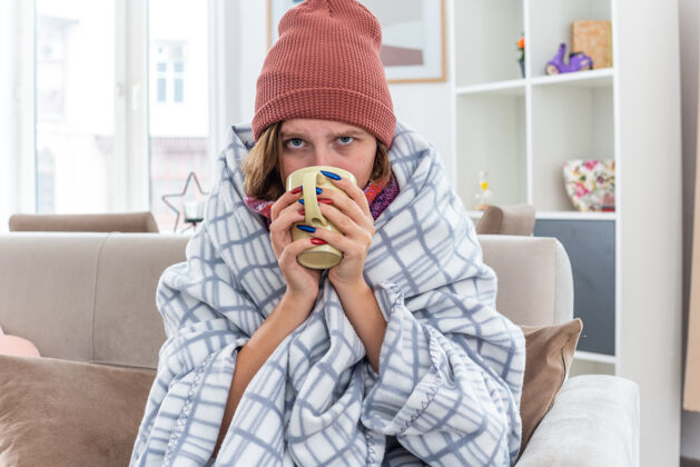女人不健康的年轻女子 戴着帽子 裹着毯子 感觉不舒服 生病 患感冒和流感 喝着热茶 坐在明亮的客厅沙发上 感觉更好喝酒包裹坐