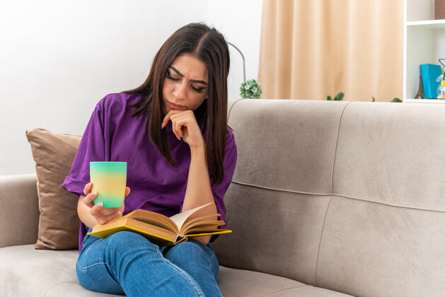 休闲一个穿着休闲服的年轻女孩 端着一杯茶 严肃地坐在客厅的沙发上看书年轻抱沙发