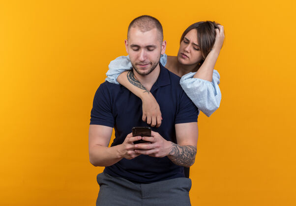 情侣一对年轻漂亮的情侣站在男友身后 怀疑地看着他的智能手机站着手机多疑