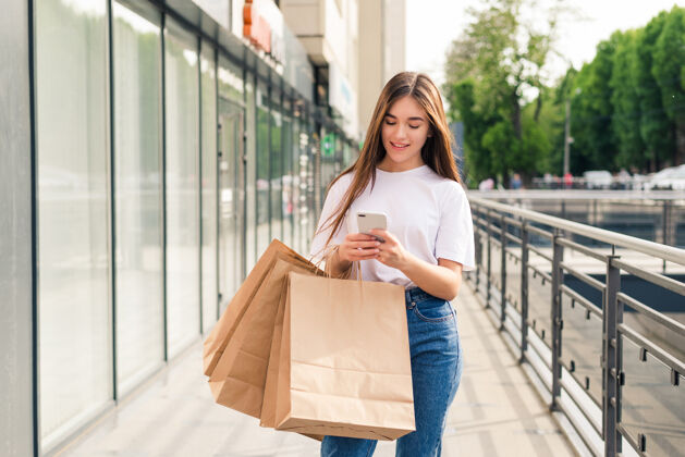 女人与朋友分享好消息美丽的年轻微笑的女人拿着购物袋和手机站在户外的特写镜头通信零售客户