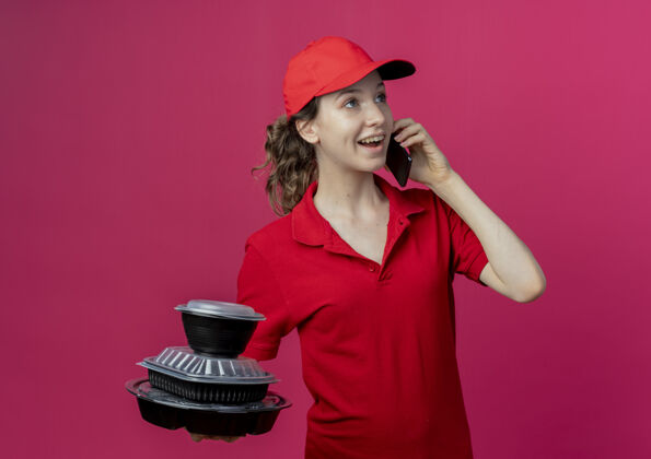 壁板快乐的年轻漂亮的送货女孩穿着红色制服 戴着帽子在打电话 拿着食物容器看着深红色空间的一边手持食物穿
