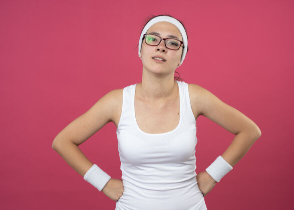 自信自信的年轻运动女孩戴着眼镜戴着头带和腕带把拳头放在腰上看着相机年轻运动粉色