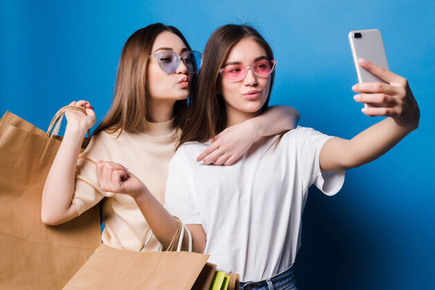 女人两个年轻的女人在电话里自拍 蓝色的墙上隔着彩色的纸袋商店销售的概念概念微笑孩子