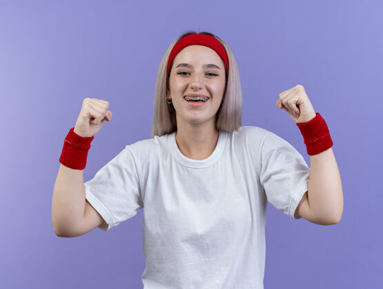穿快乐的年轻白人运动女孩戴着牙套戴着头带和手腕保持拳头腕带运动背带