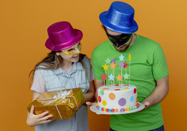 橙色惊讶的年轻女孩戴着粉色帽子和化装眼罩拿着礼盒 看着戴着化装眼罩的蓝色帽子的帅哥拿着生日蛋糕盒子礼物化装舞会