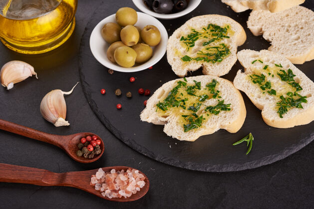 碗地中海小吃集橄榄 油 香草和切片西巴塔面包在黑色板岩石板上涂深蓝色的表面 俯视图平放大蒜小吃素食者