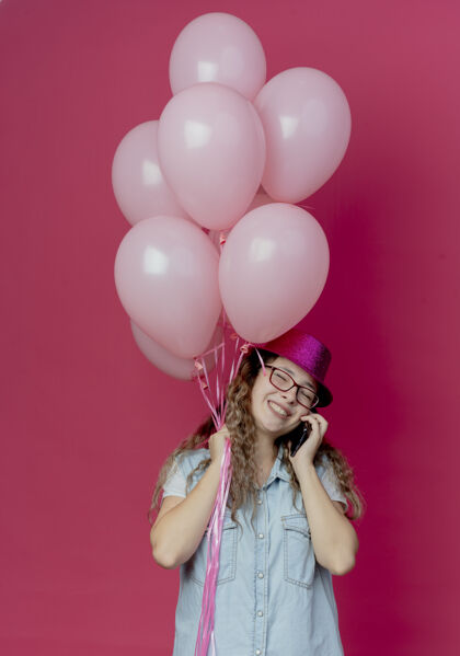 戴着一个戴着眼镜 戴着粉红色帽子 抱着气球 面带微笑的年轻女孩在电话里讲话粉色年轻封闭